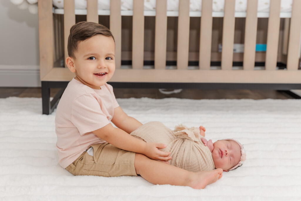 big brother sits on nursery floor in front of crib with new baby sister for newborn photos